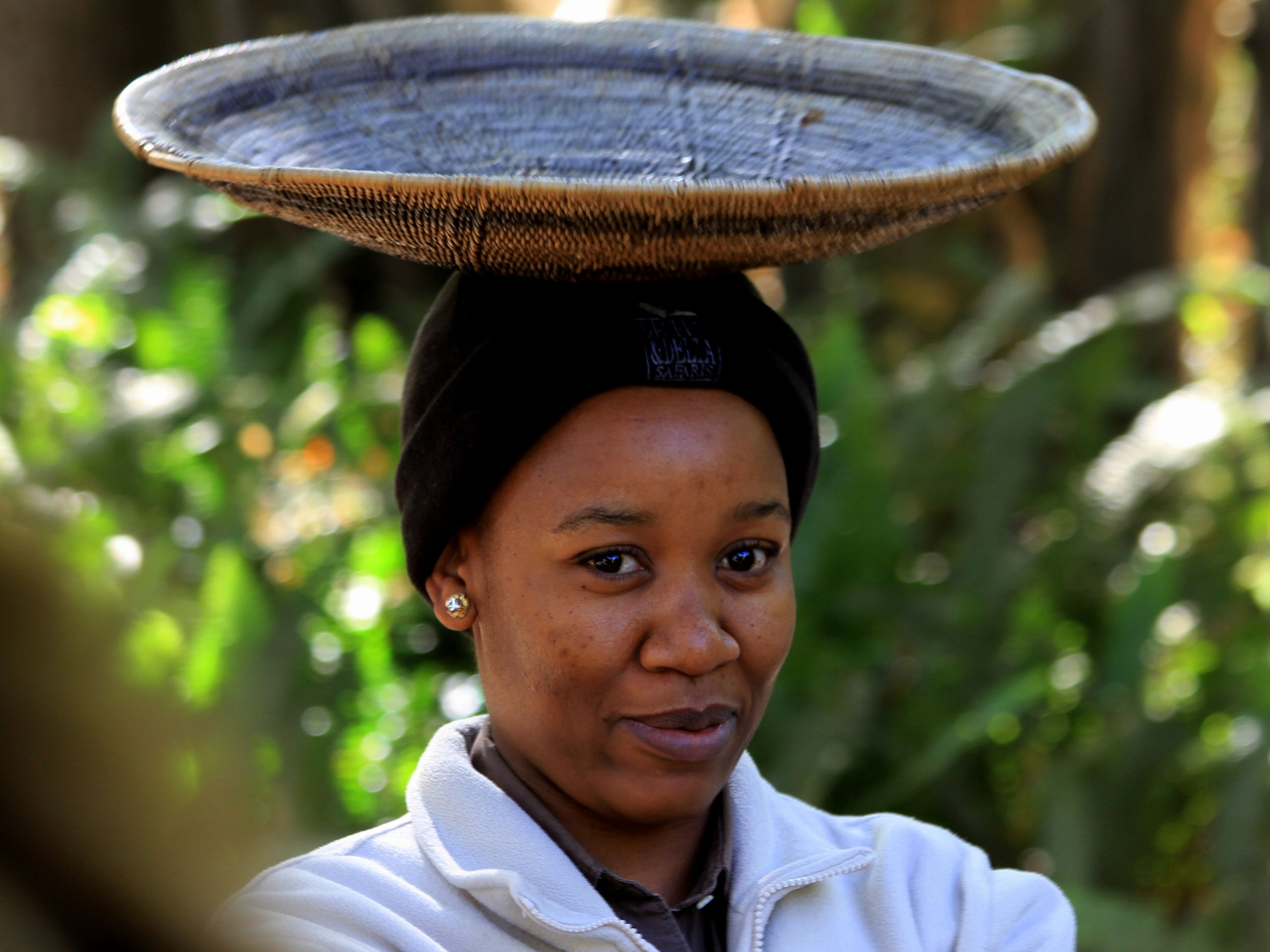 Portrait of a woman balancing a wicker tray on her head in a natural outdoor setting.